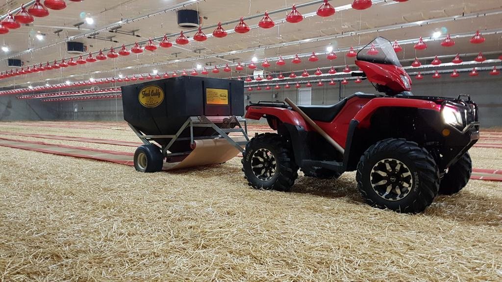A Feed Caddy hopper hitched behind a red quad in a broiler barn